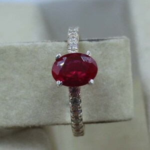 Könnte beinhalten: Ein silberner Ring mit einem großen, oval geschliffenen Rubin. Der Rubin hat eine tiefrote Farbe und ist der Mittelpunkt des Rings. Der Ring ist mit kleinen, klaren Edelsteinen besetzt. Der Ring wird vor einem neutralen Hintergrund präsentiert.