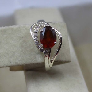 May include: A silver ring with a large, oval-cut, deep red gemstone. The gemstone is the focal point, surrounded by smaller, clear accent stones. The ring's band has a decorative, flowing design.