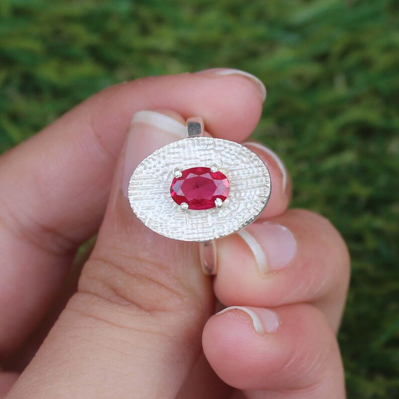 May include: A silver ring featuring an oval, textured setting with a vibrant red gemstone at its center. The ring's design is simple yet elegant, showcasing the gemstone's rich color against the silver. The ring is held against a blurred green background.