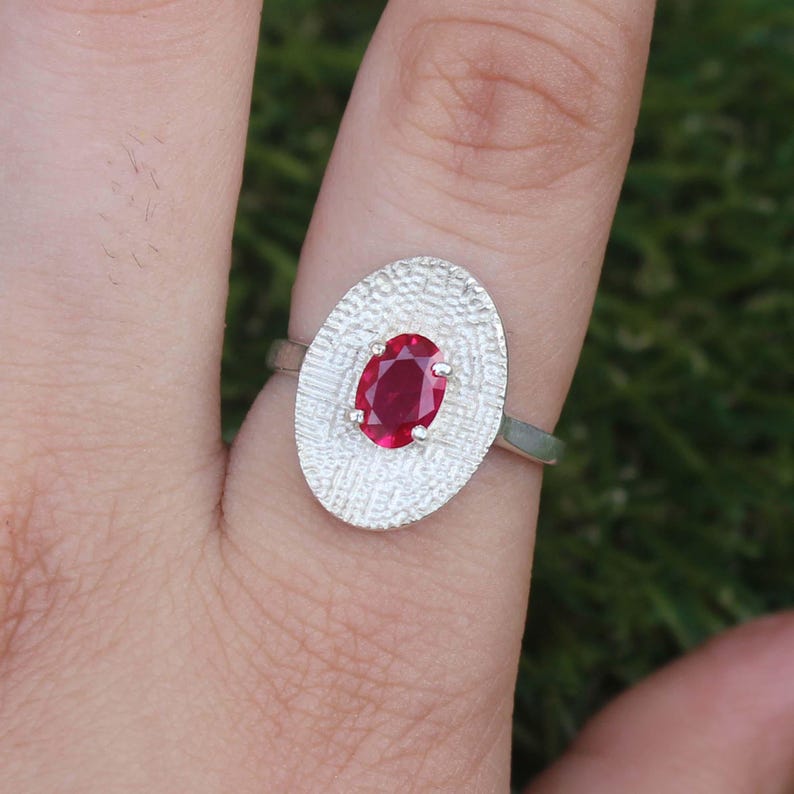 May include: A silver ring featuring an oval, textured setting with a vibrant red gemstone. The ring's band is a simple, polished silver, and the gemstone is securely held in place by small prongs. The ring is shown on a finger against a blurred green background.