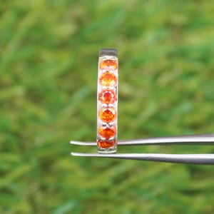 May include: A silver ring set with a line of orange gemstones. The ring is held by tweezers against a blurred green backdrop. The gemstones are evenly spaced and appear to be round cut.