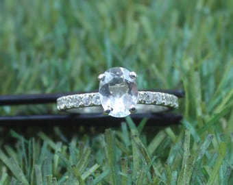 May include: A silver ring with a large, oval, light-colored gemstone as the centerpiece. The band is encrusted with smaller, clear stones. The ring is set against a backdrop of green grass, highlighting its sparkle and design.