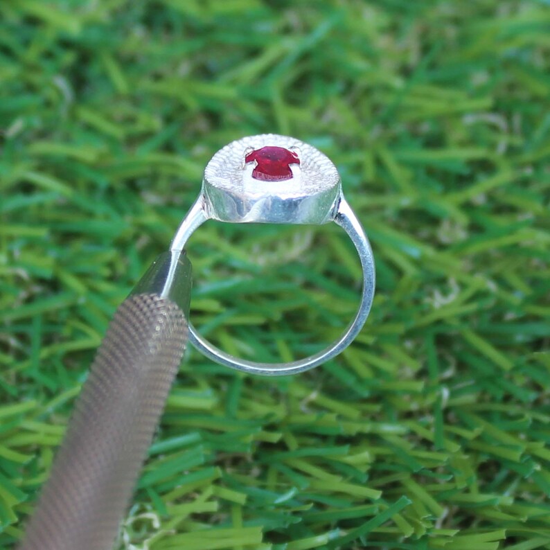 May include: A silver ring with a round, textured setting and a small, red gemstone. The ring is set against a backdrop of green grass.