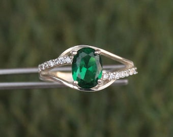 May include: A silver ring featuring a large, oval-cut emerald green gemstone. The band splits on either side of the central stone, with small, clear accent stones set along one side. The ring is held by a pair of metal tweezers against a blurred green background.
