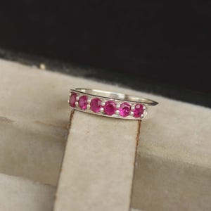 May include: A silver ring featuring a row of six oval-cut, vibrant red gemstones. The gemstones are set closely together, creating a continuous band of color. The ring is displayed against a neutral background, highlighting the sparkle of the stones.