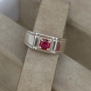 Può includere: Un anello d'argento con una vivace gemma rossa rotonda incastonata in un design rettangolare. L'anello ha una finitura lucida e uno stile classico e sobrio. La gemma è il punto focale, aggiungendo un tocco di colore.