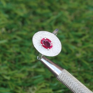 May include: A silver ring featuring an oval, deep red gemstone set in a textured, oval-shaped setting. The ring is held by a silver tool against a blurred green background.