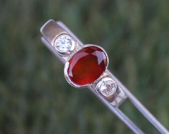 Puede incluir: Anillo de plata con una gema roja ovalada en el centro, flanqueada por dos gemas claras y redondas más pequeñas. La banda del anillo es de plata pulida. Las gemas están engastadas.