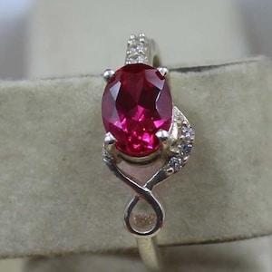 Puede incluir: Un anillo de plata con una gran gema roja oscura de corte ovalado. La gema es el punto focal, sujeta por cuatro garras. La banda del anillo tiene un diseño de símbolo de infinito con pequeñas piedras de acento en los lados.