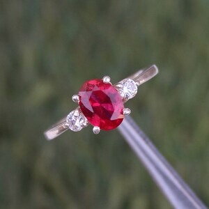 May include: A gold ring with a large oval-cut ruby centre stone, and two smaller round diamonds. The ruby is a deep red colour, and the diamonds sparkle. The ring is set against a blurred green background.