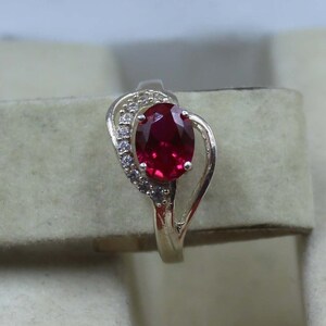 Könnte beinhalten: Ein silberner Ring mit einem großen, oval geschliffenen, tiefroten Edelstein. Der Edelstein ist von kleineren, klaren Akzentsteinen umgeben. Der Ring hat ein dekoratives, wirbelndes Design auf einer Seite.