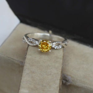 May include: A silver ring with a vibrant yellow gemstone at its centre. The band has a twisted design, embellished with small, clear accent stones. The ring is displayed in a dark box, accentuating its sparkle and elegance.
