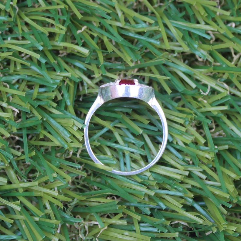 May include: A silver ring with a rectangular, dark red gemstone at the center. The gemstone is surrounded by smaller, clear stones. The ring is set against a backdrop of green artificial grass.