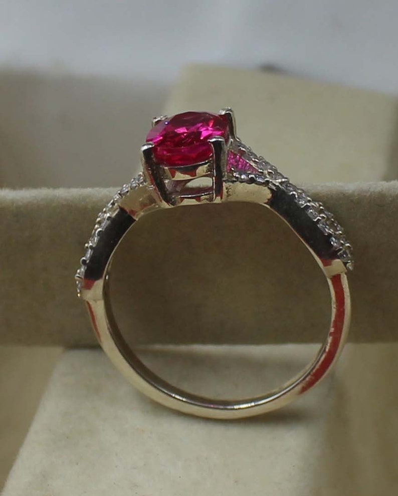 May include: A gold ring with a large, oval-cut, bright pink gemstone. The band is embellished with smaller, clear gemstones. The ring's design is showcased against a neutral backdrop. The ring's design is highlighted against a neutral backdrop.