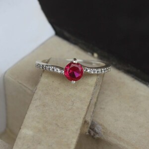 Peut inclure: Une bague en argent avec une pierre précieuse rouge vif au centre. La bande est ornée de petites pierres claires. La bague est présentée sur un fond neutre, soulignant son design élégant. Un bijou.