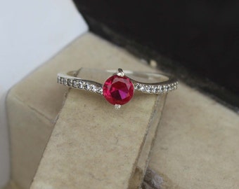 May include: A silver ring featuring a striking red gemstone at its centre. The band is embellished with small, clear stones. The ring is displayed against a neutral backdrop, accentuating its elegant design. A piece of jewellery.