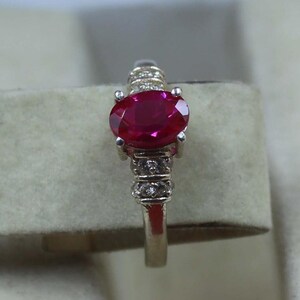 Peut inclure: Une bague en argent ornée d'un grand rubis de forme ovale en son centre. Le rubis est d'un rouge vif. La monture de la bague est ornée de petits diamants transparents. La bague est posée sur un fond neutre.