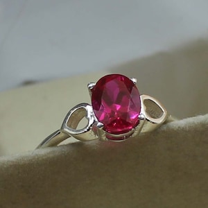 Może przedstawiać: Srebrny pierścionek z dużym, owalnym, jasno czerwoną kamieniem szlachetnym. Pierścionek ma delikatną obrączkę i dekoracyjne ażurowe akcenty po obu stronach kamienia szlachetnego, tworząc unikalny i elegancki design. Pierścionek jest na neutralnym tle.