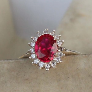 Puede incluir: Un anillo con una gran piedra preciosa de color rojo intenso y corte ovalado en el centro, rodeada por un halo de pequeñas piedras claras de corte redondo. El engaste es una banda metálica que sostiene el conjunto en una forma simétrica.
