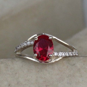 Könnte beinhalten: Ein silberner Ring mit einem großen, oval geschliffenen, tiefroten Edelstein. Das Band teilt sich auf beiden Seiten des zentralen Steins, mit kleinen, klaren Akzentsteinen, die entlang einer Seite jeder Spaltung gesetzt sind. Schmuck.