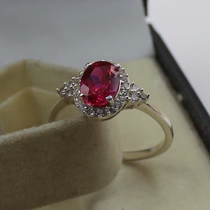 Puede incluir: Un anillo de plata con un gran rubí de corte ovalado como pieza central. El rubí está rodeado por un halo de piedras preciosas más pequeñas y transparentes. Piedras preciosas transparentes adicionales están engastadas a ambos lados de la piedra principal, creando un diseño elegante y brillante.