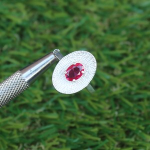 May include: A silver ring with an oval, deep red gemstone set in a textured, oval silver setting. The ring is displayed against a green background, with a silver tool in the frame.