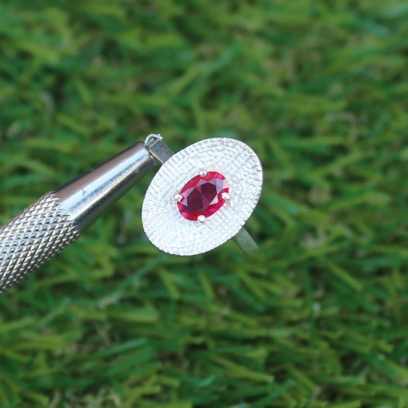 May include: A silver ring with an oval, deep red gemstone set in a textured, oval silver setting. The ring is displayed against a green background, with a silver tool in the frame.