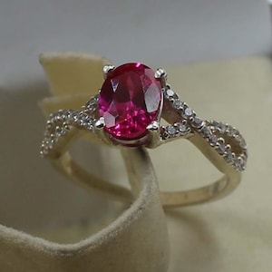 May include: A gold-toned ring featuring a large, oval-cut, vibrant red gemstone as the centerpiece. The band splits into a delicate, intertwined design, embellished with small, sparkling accent stones, creating an elegant and eye-catching piece of jewelry.