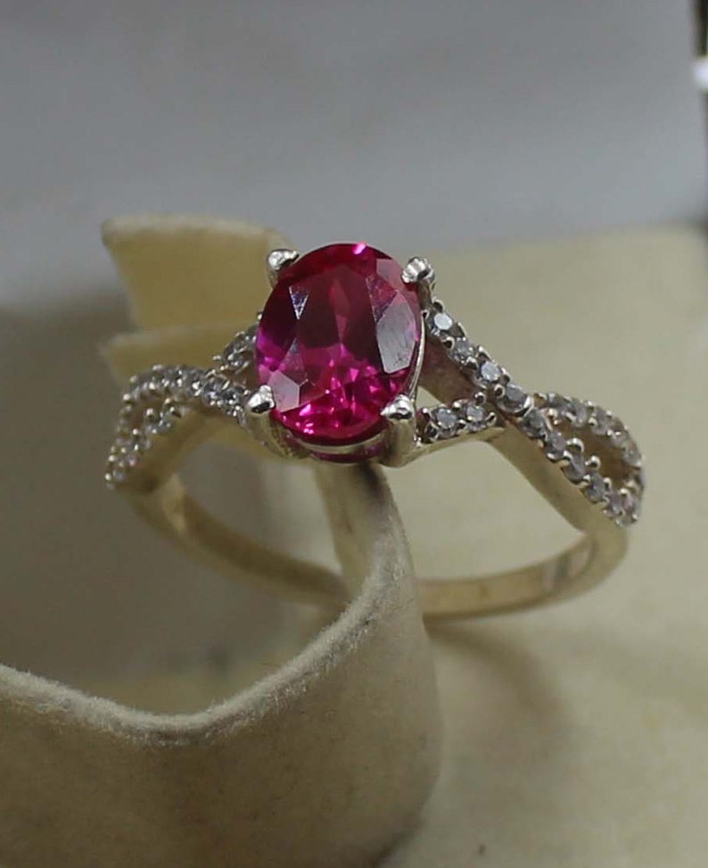 May include: A gold ring with a large, oval-cut, bright red gemstone at its centre. The band splits into a delicate, intertwined design, adorned with small, sparkling accent stones, creating an elegant and eye-catching piece of jewellery.
