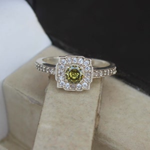 May include: A silver ring featuring a square halo design. The center stone is a round, olive-green gem, surrounded by a square halo of white stones. The band is also adorned with small white stones, creating a sparkling effect.