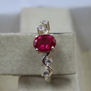 Könnte beinhalten: Ein silberner Ring mit einem großen, oval geschliffenen Rubin. Der Rubin hat eine leuchtend rote Farbe. Der Ring ist mit kleineren, klaren Edelsteinen verziert. Das Design hat eine fließende, organische Form.