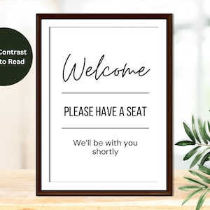 May include: A black and white sign with a brown frame that reads "Welcome, Please Have a Seat, We'll be with you shortly."