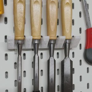 May include: Four wood chisels with different blade widths (8mm, 12mm, 18mm, and 25mm) are displayed on a white pegboard. The chisels have light-coloured wooden handles and silver metal blades, suitable for woodworking.