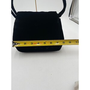 May include: A black velvet handbag with a flap closure and a cord strap. The bag is approximately 7 inches wide, as measured by a yellow tape measure. The bag has a small decorative pattern near the bottom.