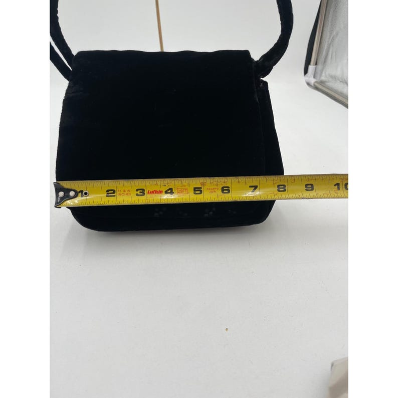May include: A black velvet handbag with a flap closure and a cord strap. The bag is approximately 7 inches wide, as measured by a yellow tape measure. The bag has a small decorative pattern near the bottom.