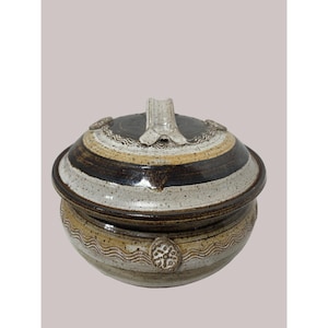May include: A handmade ceramic lidded bowl with a rustic design. The bowl features a dark brown, white, and tan striped lid with a handle. The base of the bowl has a wavy pattern and a decorative emblem. The pottery is a neutral color.
