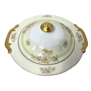 Puede incluir: Fuente para servir de color crema con tapa. La fuente presenta un asa dorada a cada lado y un pomo dorado en la tapa. La tapa y la fuente están decoradas con motivos florales en rosa, amarillo y verde.
