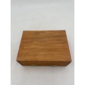 May include: A rectangular wooden box with a hinged lid. The box is made of light brown wood with visible grain and brass-colored hinges. The box is closed and sits on a white surface.