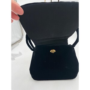 May include: A small, black velvet handbag with a gold-colored clasp. The bag has a structured shape with a flap closure and two black cord handles. The interior of the bag is also black.