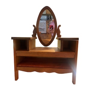 May include: A wooden vanity with an oval mirror and two side shelves. The vanity has a lower shelf and a scalloped edge. The wood has a warm, natural finish, and the mirror is framed in the same wood.