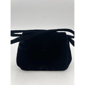 May include: A small, black velvet crossbody bag with a rectangular shape and a flap closure. The bag has a long, thin strap for over-the-shoulder or crossbody wear. The velvet material gives the bag a soft, luxurious appearance.