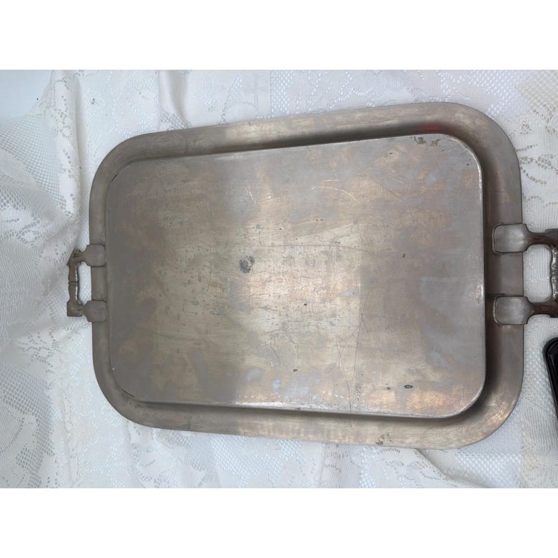 May include: A rectangular metal serving tray with a muted brass-toned finish and a smooth, flat central surface. The tray features rounded corners and two sturdy, integrated handles on the shorter sides, showing signs of surface wear.