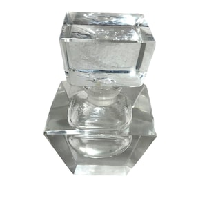 May include: A clear, faceted glass perfume bottle with a square stopper. The bottle has a geometric design with a hexagonal base and a smaller, rounded body. The stopper is also square and faceted.