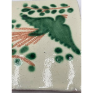 May include: Square ceramic tile with a hand-painted design. The tile features a green bird with a branch and berries in shades of green and orange against a cream background. The tile has a rustic, handmade appearance.