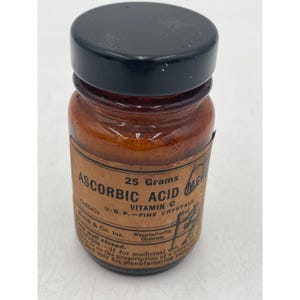 May include: A vintage brown glass bottle with a black lid, labeled "ASCORBIC ACID" and "25 Grams." The label also indicates "Vitamin C" and "U.S.P. - FINE CRYSTALS." The bottle is from Merck & Co. Inc.