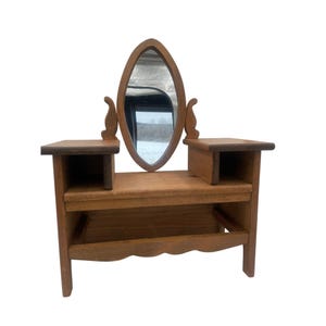 May include: A small, wooden vanity with an oval mirror. The vanity has two side shelves and a lower shelf. The mirror is framed by the wood. The wood has a natural finish. The vanity is a vintage style.