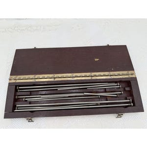 Puede incluir: Una caja de almacenamiento rectangular de madera marrón oscuro con una bisagra de metal color latón y dos cierres frontales. Dentro de la caja abierta hay varias varillas metálicas cilíndricas de color plateado dispuestas horizontalmente.