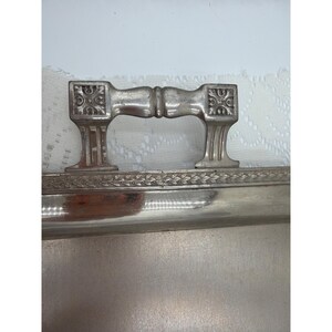 May include: A rectangular silver-toned metal tray featuring a polished surface and a decorative border with a repeating leaf pattern. The tray includes a sturdy, ornate metal handle with square ends and embossed floral details on each side.