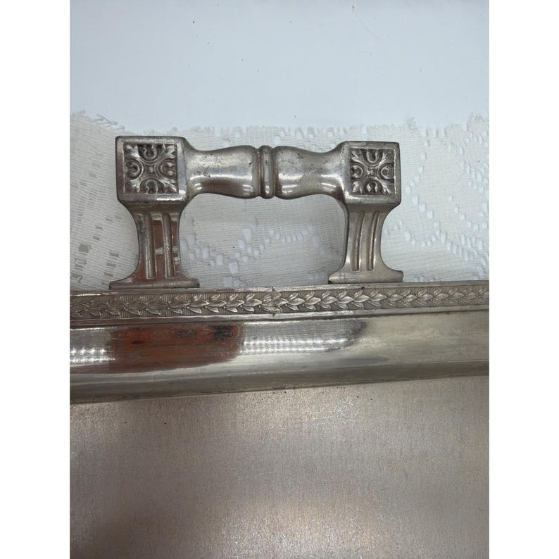 May include: A rectangular silver-toned metal tray featuring a polished surface and a decorative border with a repeating leaf pattern. The tray includes a sturdy, ornate metal handle with square ends and embossed floral details on each side.