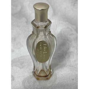 May include: A small, clear glass perfume bottle with a gold-colored cap and a gold label that reads "Estee Lauder Skin Perfume". The bottle has a decorative, fluted design and a slightly amber hue at the base.